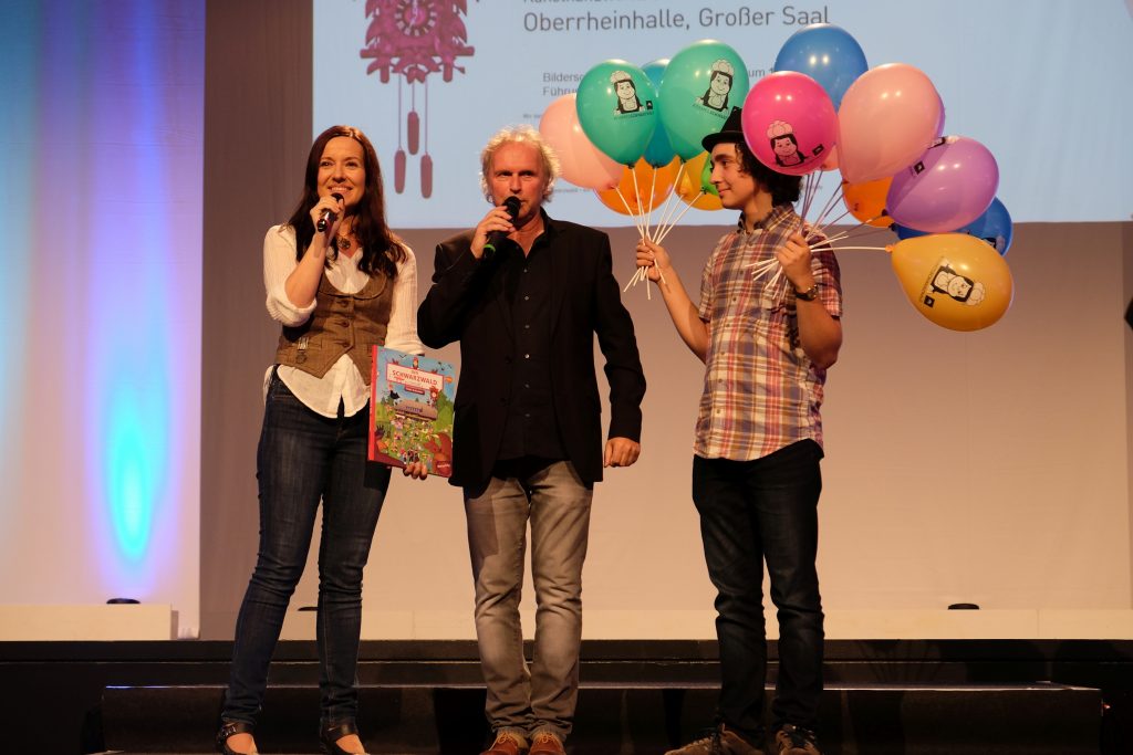 Das Bühnenbild zeigt links Katja Schneider mit Miktofon, mittig Uwe Baumann mit Mikrofon und rechts einen jungen Mann mit bunten Anni-Luftballons