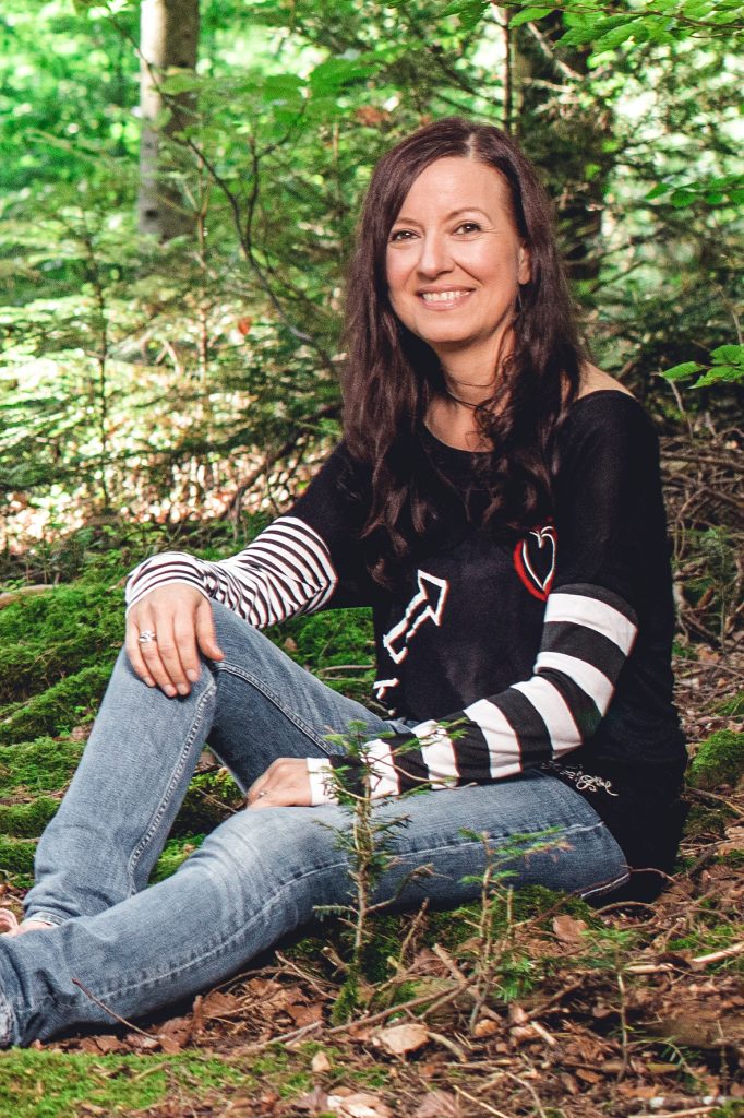 Katja Schneider sitzt mit einem ausgestreckten und einem halb angewinkelten Bein im Wald und schaut mit seitlichem Blick in die Kamera
