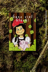 Auf der Postkarte steht "Ganz viel Glück" und zeigt Anni vor einem Holzbrett mit einem Glücksschweinchen in der Hand und vielen Kleeblättern rund die beiden herum