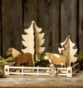 Auf einer massiven Holzplatte steht das Anni-Holzspiel. Dieses besteht aus einem Zaun, dem Pferd Mecki, der Kuh Elsa und zwei Tannenbäumen